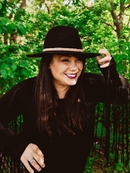 Photographer Kendra Pierroz in a wide-brimmed hat smiling at the camera in front of dense foliage.