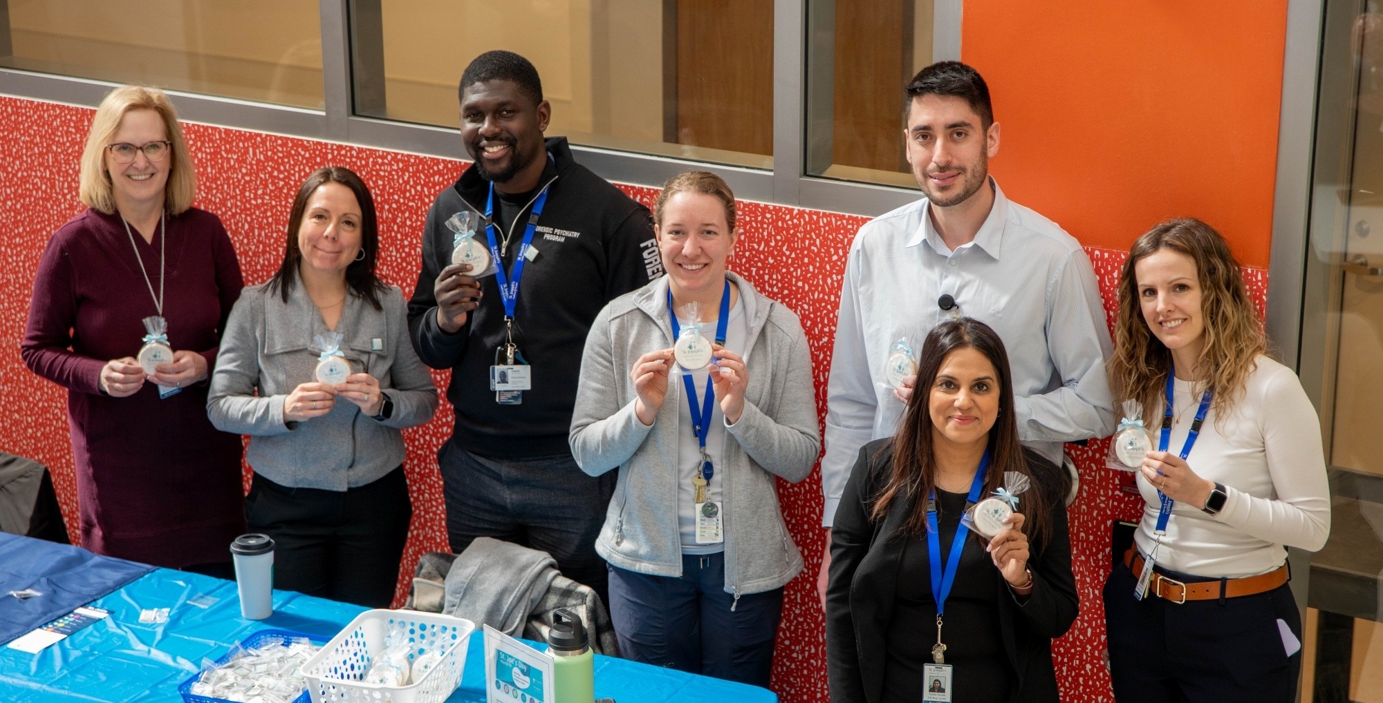 A group of 7 staff members gather together at a table, holding up small bags. They look up to the camera and smile.
