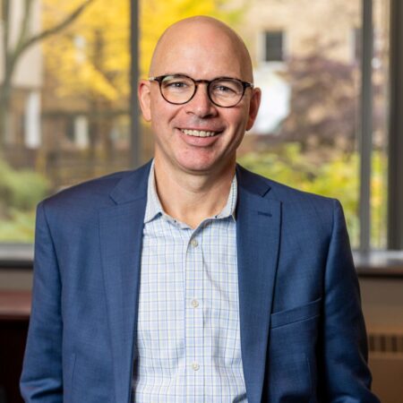 Dr. Heenan smiles at the camera. He has a shaved head and light-toned skin. He wears glasses, and a navy blue suit jacket.
