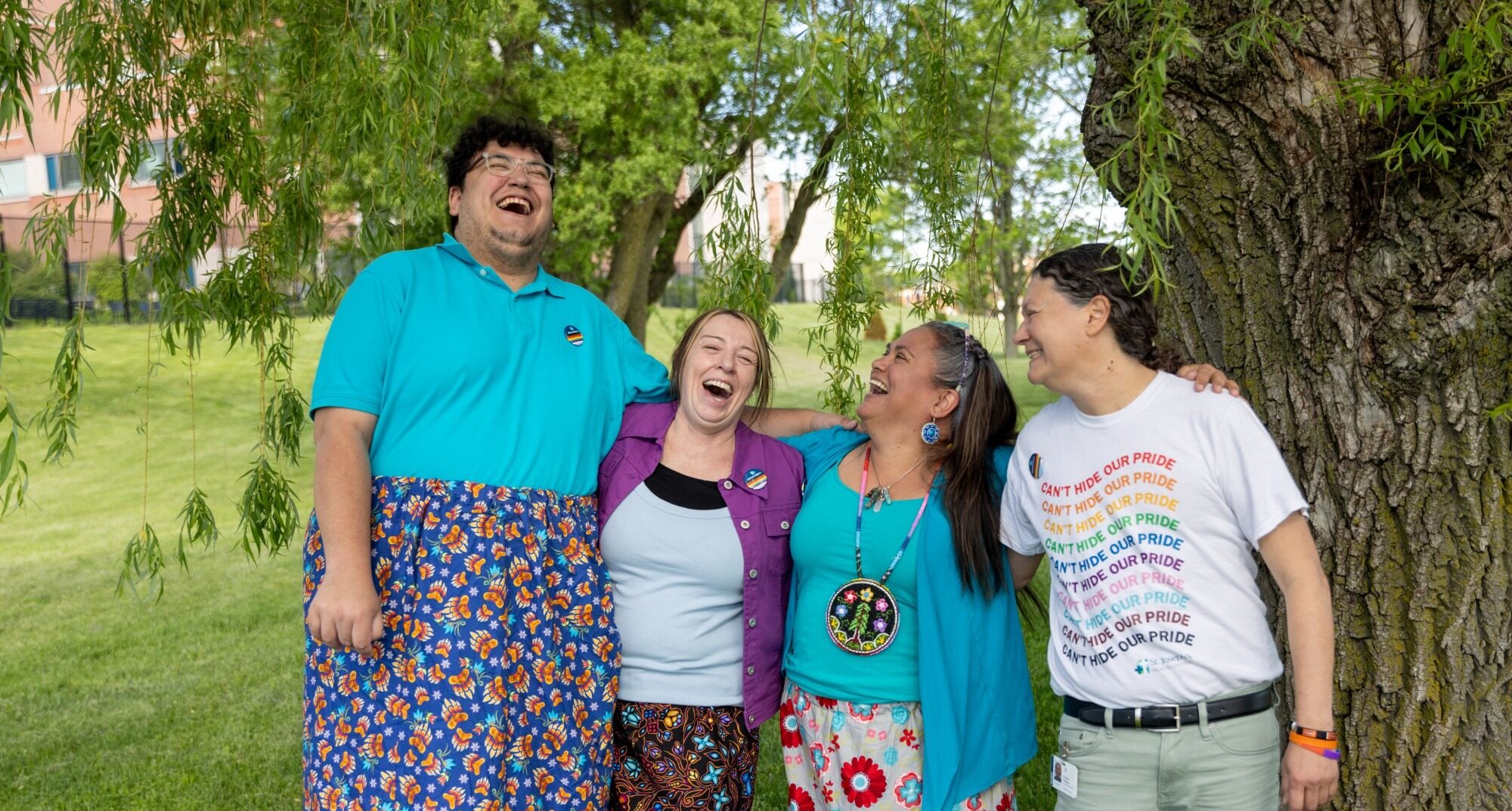 4 people stand together near a tree with their arms around each other while laughing. 3 wear bright patterned colourful skirts, 1 wears a 