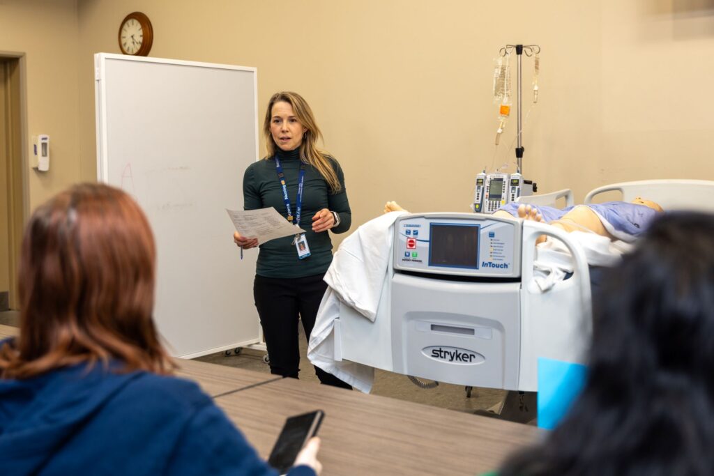 A staff member stands in a staged medical room with a bed and a medical mannequin. She addresses attendees sitting in the room.