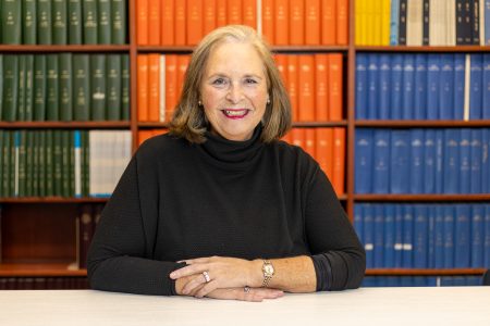 Barbara sits in front of a shelf full of books with her hands stacked together. She faces the camera smiling. She has light-toned skin, shoulder-length blonde hair, and wears a black turtleneck.